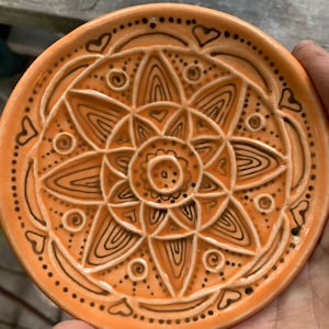 Puede incluir: Un plato redondo de cerámica naranja con un diseño de mandala negro. El mandala presenta detalles intrincados y un patrón repetitivo de corazones.