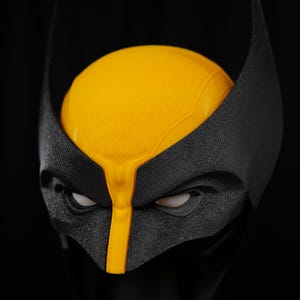 Puede incluir: Una máscara de Wolverine negra y amarilla, con una superficie negra texturizada y detalles amarillos. La máscara tiene orejas puntiagudas y aberturas blancas para los ojos. La sección amarilla cubre la parte superior de la cabeza y el puente de la nariz.