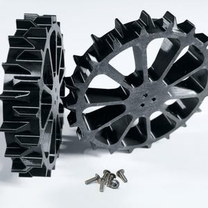 Könnte beinhalten: Zwei schwarze, zahnradförmige Komponenten mit spitzen Zähnen, vor weißem Hintergrund. Mehrere kleine silberne Schrauben sind in der Nähe der Zahnräder verstreut. Die Zahnräder haben ein komplexes, offenes Design mit mehreren Speichen.