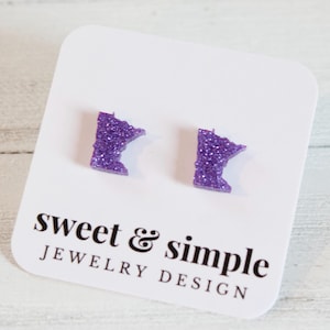 May include: A pair of purple glitter stud earrings in the shape of the state of Minnesota. The earrings are displayed on a white card with the text "sweet & simple JEWELRY DESIGN" in black.