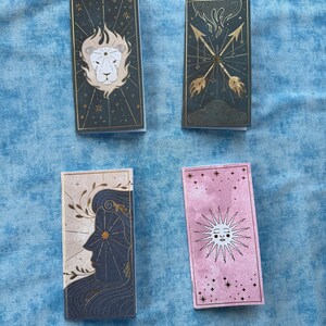 May include: Four rectangular tarot card decks. The decks feature illustrations in gold, white, and blue on a dark green, pink, and beige background. The designs include a lion, arrows, a profile, and a sun with stars.