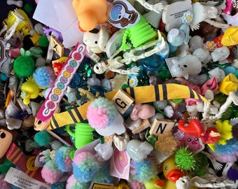 Puede incluir: Un colorido surtido de pequeños objetos decorativos, que incluyen pompones, figuras de esqueletos y bloques de letras de madera. Se ven varios colores y texturas, creando una mezcla lúdica y ecléctica. Los artículos parecen ser materiales de manualidades o elementos decorativos.
