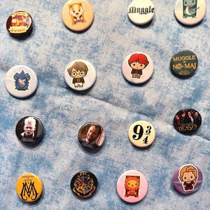 May include: A collection of round, colorful pinback buttons featuring various designs and characters. Some buttons display text like "Muggle" and "9 3/4", while others showcase illustrations of cartoon characters and logos. The buttons are arranged on a light blue surface.