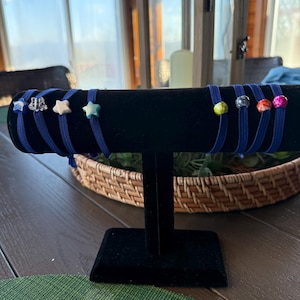 May include: A display of blue elastic hair ties, each adorned with a decorative bead. The beads include star and flower shapes, as well as colorful round beads. The ties are arranged on a black velvet display stand.