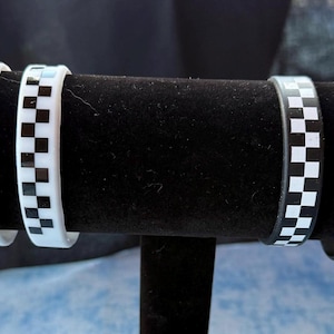 May include: A display of several bracelets with a black and white checkered pattern. The bracelets are arranged on a black velvet display stand. The bracelets are made of a glossy material.