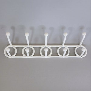 May include: A white metal wall-mounted coat rack with five hooks. Each hook is a rounded knob on a slender stem, connected to a series of circular designs. The rack is mounted on a gray wall.