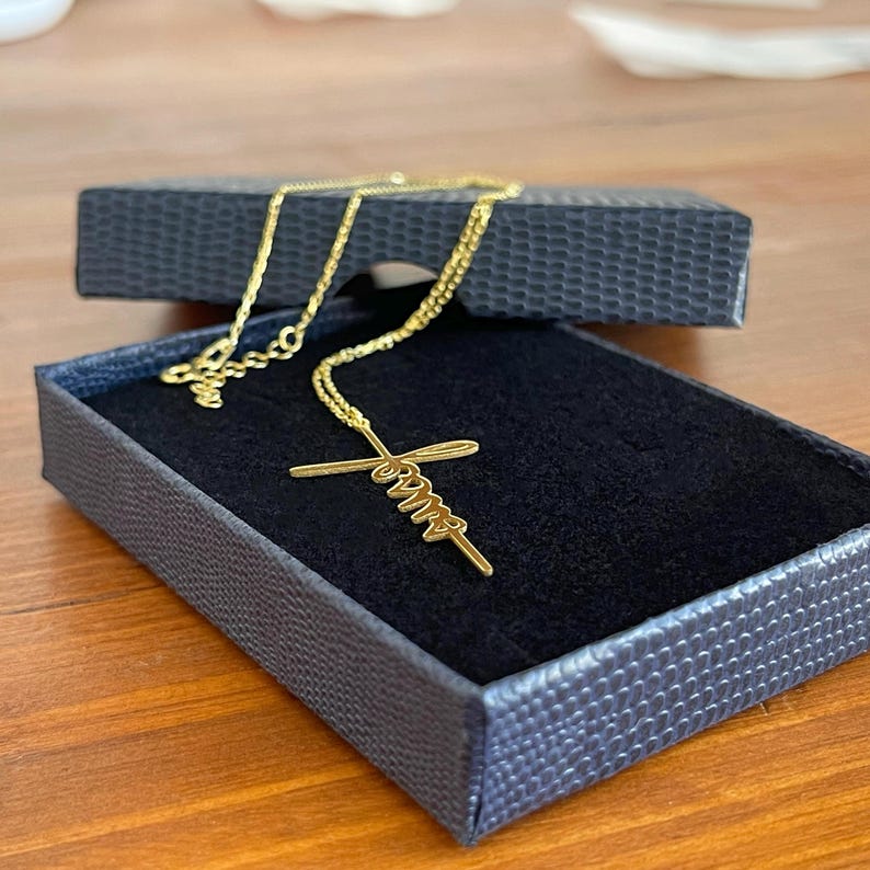 K&ouml;nnte beinhalten: Eine goldfarbene Halskette mit Kreuzanh&auml;nger, pr&auml;sentiert in einer schwarzen Schmuckschatulle. Der Anh&auml;nger tr&auml;gt das Wort "Faith" in Schreibschrift. Die zarte Kette ist vor dem dunklen Samt der Schatulle sichtbar.