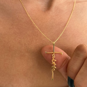 K&ouml;nnte beinhalten: Eine goldfarbene Halskette mit einem Kreuzanh&auml;nger. Das Wort "faith" ist vertikal auf dem Kreuz geschrieben. Die Halskette hat eine feine Kette. Der Hintergrund ist eine verschwommene Hautansicht.