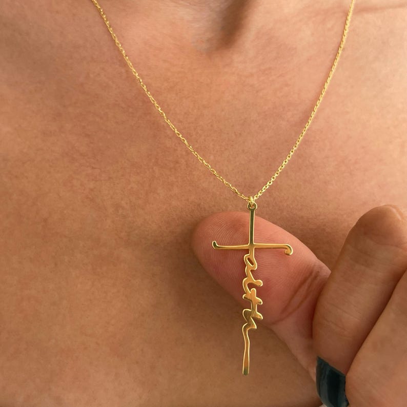 K&ouml;nnte beinhalten: Eine goldfarbene Halskette mit einem Kreuzanh&auml;nger. Das Wort "faith" ist vertikal auf dem Kreuz geschrieben. Die Halskette hat eine feine Kette. Der Hintergrund ist eine verschwommene Hautansicht.