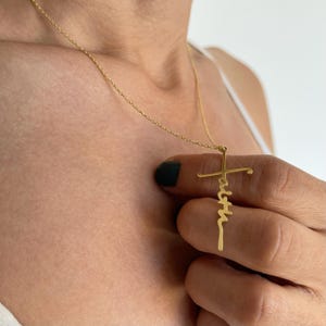 K&ouml;nnte beinhalten: Eine goldfarbene Halskette mit einem Kreuzanh&auml;nger. Das Wort "faith" ist vertikal auf dem Kreuz geschrieben. Die Halskette wird um den Hals getragen.