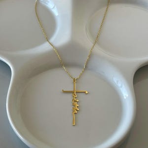 K&ouml;nnte beinhalten: Eine goldfarbene Halskette mit einem Kreuzanh&auml;nger und dem Wort "faith" vertikal geschrieben. Die Halskette wird auf einer wei&szlig;en, geriffelten Schale pr&auml;sentiert. Kette und Anh&auml;nger sind aus gl&auml;nzendem Metall.