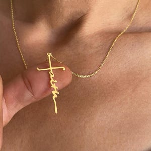 K&ouml;nnte beinhalten: Eine goldfarbene Halskette mit einem Kreuzanh&auml;nger, auf dem das Wort "faith" in Schreibschrift steht. Der Anh&auml;nger h&auml;ngt an einer zarten Kette. Die Halskette wird vor einem hautfarbenen Hintergrund gezeigt.