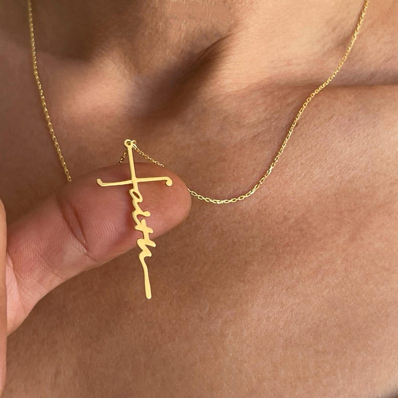 K&ouml;nnte beinhalten: Eine goldfarbene Halskette mit einem Kreuzanh&auml;nger, auf dem das Wort "faith" in Schreibschrift steht. Der Anh&auml;nger h&auml;ngt an einer zarten Kette. Die Halskette wird vor einem hautfarbenen Hintergrund gezeigt.
