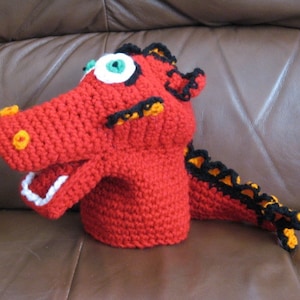 May include: Red crocheted dragon puppet with black and yellow accents. The puppet has a large mouth with white teeth and green eyes.