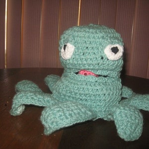 May include: A crocheted green octopus with white eyes and a pink tongue. The octopus has eight tentacles and is sitting on a wooden surface.