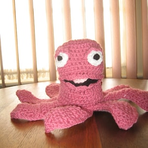 May include: A pink crocheted octopus with white eyes. The octopus is sitting on a wooden table.