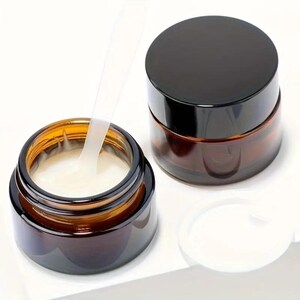May include: Two amber glass jars with black lids. One jar is filled with a white cream and a clear plastic spatula is inside the jar. The other jar is empty.