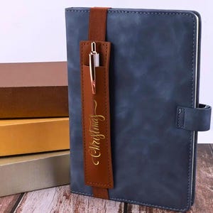 May include: A blue faux leather journal with a brown leather pen holder and elastic closure. The pen holder has the word "Christmas" written in gold lettering.