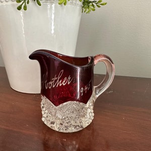 以下が含まれることがあります： クリアなカットガラスのベースを持つ、ルビーレッドの小さなガラス製のクリーマー。クリーマーにはハンドルが付いており、「Mother」と「945」という文字がエッチングされています。
