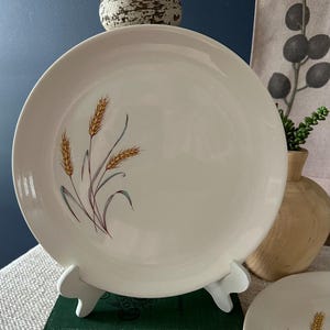 Puede incluir: Plato de cerámica blanquecina con un diseño de espigas de trigo en tonos dorados, morados y azules. El plato se exhibe sobre un soporte blanco. Se ve parcialmente otro plato con el mismo diseño.