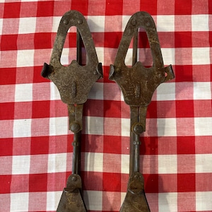 May include: Two antique, rusty metal ice tongs. Each tong has a triangular handle, a central bar, and a hinged lower section with a curved, claw-like design. The tongs are displayed on a red and white checkered tablecloth.