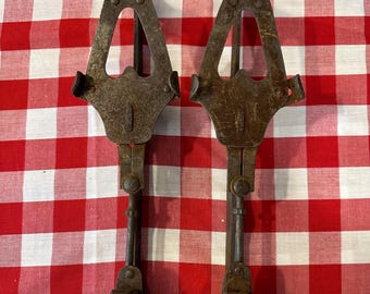 May include: A pair of antique, rusty metal ice tongs. Each tong features a triangular handle, a central bar, and a hinged lower section with a curved, claw-like design. The tongs are set against a red and white checked tablecloth.