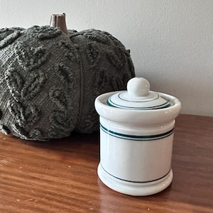 May include: A white ceramic sugar bowl with a matching lid, featuring thin dark green horizontal stripes around the body and the lid. The container has a cylindrical shape with a small knob handle on top of the lid and a smooth finish.
