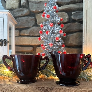 May include: A ruby red glass creamer and sugar bowl set with handles. The set is on a wooden surface with a small silver Christmas tree in the background. The creamer has a spout and the sugar bowl has two handles.