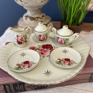 Puede incluir: Un juego de té de porcelana vintage. La bandeja color crema contiene una lechera, un azucarero, una tetera y dos platillos. Cada pieza está decorada con rosas rojas y rosas y detalles dorados. La bandeja tiene un borde festoneado y detalles de estrella.