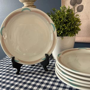 May include: A set of off-white ceramic plates with a decorative border of light brown and teal arcs. The plates are displayed on a black stand and stacked on a blue and white checkered tablecloth. A small potted plant is in the background.