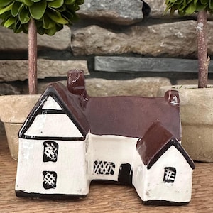 May include: A ceramic miniature house with a white exterior, brown roof, and chimney. The house features black window and door details. The design is reminiscent of a quaint cottage, perfect for home decor or a collector's item.