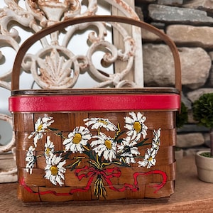 Könnte beinhalten: Ein Vintage-Picknickkorb mit Holzgriff und rotem Rand. Der geflochtene Korb hat ein Blumendesign aus weißen Gänseblümchen mit gelben Zentren und roten Bändern. Ein charmantes Dekorationsstück.