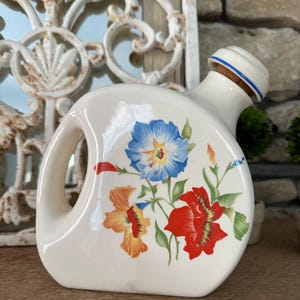 May include: White ceramic jug with a floral design featuring blue, red, orange, and green flowers. The jug has a cork stopper and a handle.