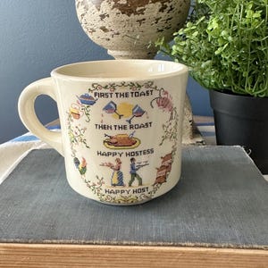 May include: Cream-colored ceramic mug with a handle, featuring cross-stitch style embroidery. The mug has the text "FIRST THE TOAST, THEN THE ROAST, HAPPY HOSTESS, HAPPY HOST" and illustrations of food and people.