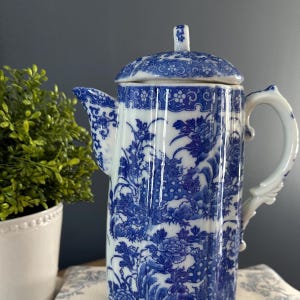 May include: A tall, white and blue porcelain coffee pot with a lid. The pot features a detailed floral and foliage pattern in blue against a white background. The handle and spout are white. The pot is a decorative item.