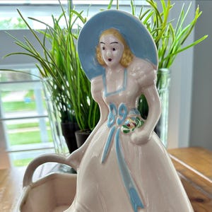 May include: A ceramic figurine planter featuring a person with blonde hair wearing a wide-brimmed light blue hat and a pale pink dress with blue trim. The figure holds a small bouquet of flowers and is attached to a hollow, square-shaped base.