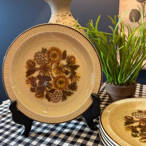 May include: Tan and brown floral patterned ceramic plates. The plates have a textured rim and a central floral design in shades of brown and yellow. The plates are displayed on a black stand, with a checkered tablecloth in the background.