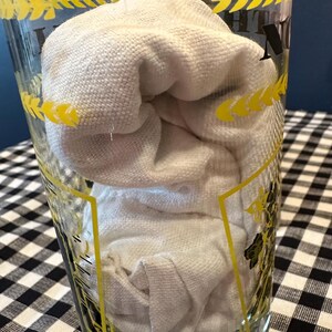 May include: A clear glass with yellow accents and a white cloth inside. The glass has a decorative pattern around the rim. The cloth is folded and fills the majority of the glass. The background is a black and white checkered pattern.