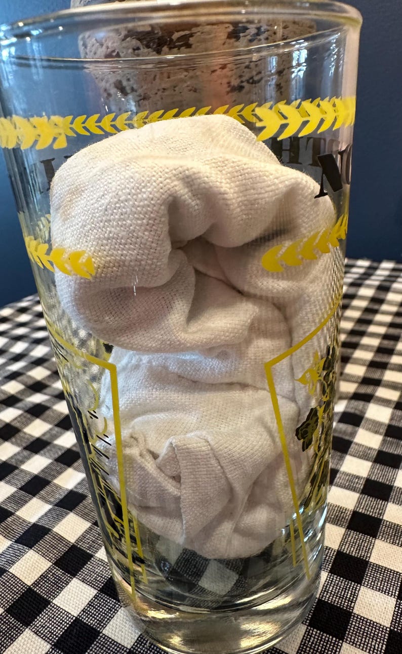 May include: A clear glass with yellow accents and a white cloth inside. The glass has a decorative pattern around the rim. The cloth is folded and fills the majority of the glass. The background is a black and white checkered pattern.