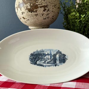 May include: Oval, off-white serving platter with a blue and white transferware design of a house, tree, and gate. The platter is on a red and white checkered tablecloth.