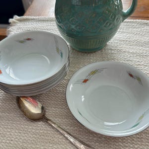 May include: A set of white ceramic bowls with a silver rim and a colorful geometric design. A silver spoon and a turquoise pitcher are also visible. The bowls are stacked and appear to be part of a dinnerware set.
