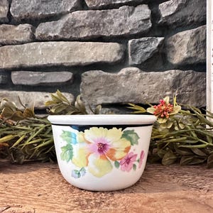 May include: A white ceramic bowl featuring a black rim and a floral design on the front. The pattern displays a large yellow and orange flower with green leaves and smaller pink blossoms painted on the exterior surface of the rounded vessel.