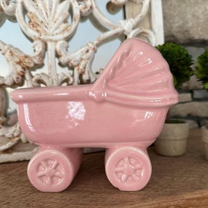 May include: A pink ceramic baby carriage planter with a hood and four wheels. The carriage has a classic design and is likely intended for decorative purposes. The wheels have a starburst design.