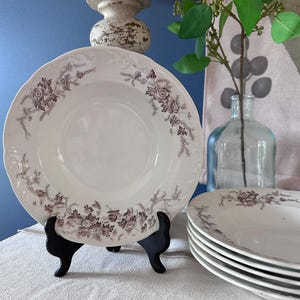 May include: A set of white ceramic soup bowls featuring a scalloped edge and a brown floral transferware pattern around the rim. One bowl is displayed upright on a black stand, while a stack of four additional matching bowls sits to the right.