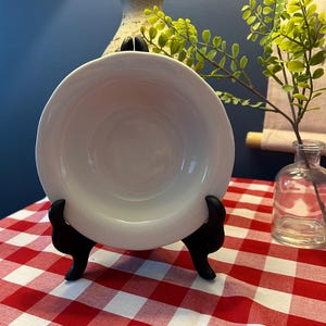 Può includere: Una ciotola in ceramica bianca esposta su un supporto nero. La ciotola ha un design semplice e arrotondato. Il supporto è posizionato su una tovaglia a scacchi rossi e bianchi. Un piccolo vaso di vetro con vegetazione è visibile sullo sfondo.