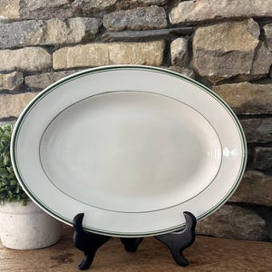 May include: An oval, white serving platter with a green border and a thin green accent line. The platter is displayed on a black stand, set against a stone wall background. A small potted plant is visible on the left.