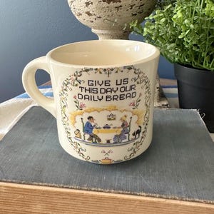 May include: Cream-colored ceramic mug with a handle, featuring a cross-stitch design. The design includes the text "GIVE US THIS DAY OUR DAILY BREAD" and a scene of two people at a table. The mug is sitting on a book.