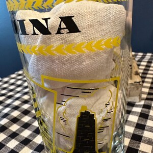May include: Clear glass with yellow accents and the word "INA" in black. The glass features a black and yellow illustration of a lighthouse. The glass is filled with a white cloth.