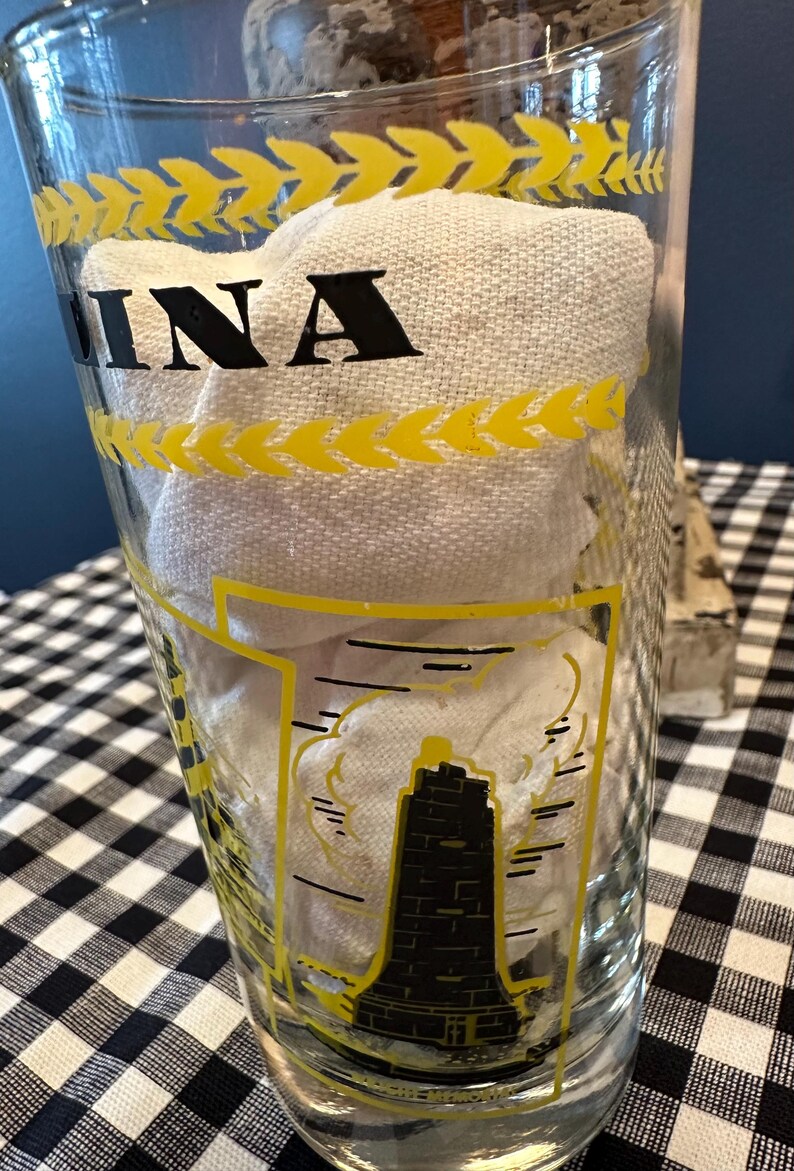 May include: Clear glass with yellow accents and the word "INA" in black. The glass features a black and yellow illustration of a lighthouse. The glass is filled with a white cloth.