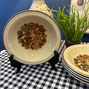 Puede incluir: Un juego de cuencos de cerámica vintage con un acabado beige moteado y un diseño floral marrón. Cada cuenco tiene un borde marrón y se exhibe sobre un soporte negro. Los cuencos están apilados sobre una superficie a cuadros en blanco y negro.